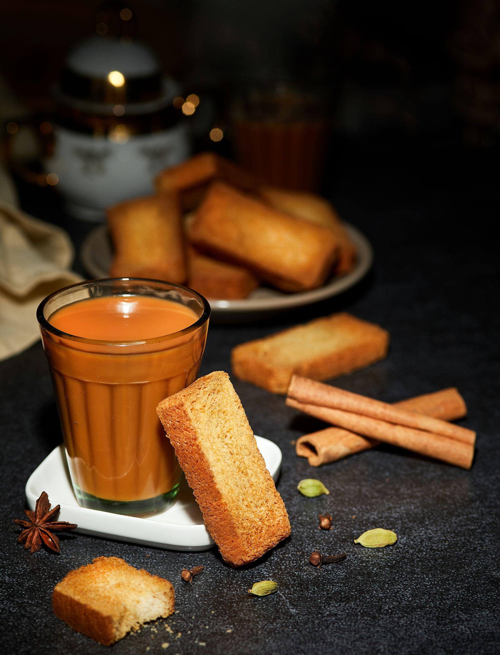 A glass of masala chai on a saucer with rusk, cinnamon, star anise and cardamom scattered around it
